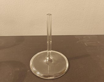 May include: A clear glass stand featuring a circular base and a slender, vertical central stem. The stem has a subtle, segmented texture resembling bamboo. The object is transparent and constructed from smooth, molded glass material.