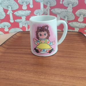 May include: A white ceramic mug featuring a colorful illustration of a Raggedy Ann doll with red yarn hair, wearing a yellow dress with floral accents. The doll is centered on a soft pink background on the front of the mug, which has a curved handle.