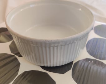 May include: A white, ribbed ceramic ramekin with a slightly textured rim. The small, round dish sits on a tray with a black and white pattern. The ramekin is empty and ready for use.