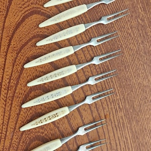 May include: A set of twelve small metal cocktail forks with two tines each. The handles are off-white with a delicate, embossed floral pattern in a light metallic tone. The forks are arranged in a diagonal line, showcasing the uniform design.