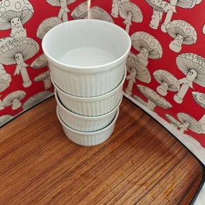 May include: A stack of four white ceramic ramekins with vertical ribbed sides. Each ramekin has a smooth interior and a consistent cylindrical shape, designed for individual servings of food. The stack is positioned upright on a flat surface.