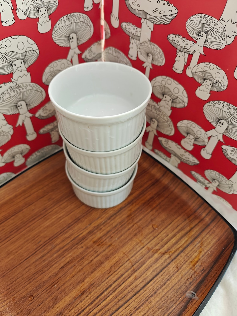 May include: A stack of four white ceramic ramekins with vertical ribbed sides. Each ramekin has a smooth interior and a consistent cylindrical shape, designed for individual servings of food. The stack is positioned upright on a flat surface.