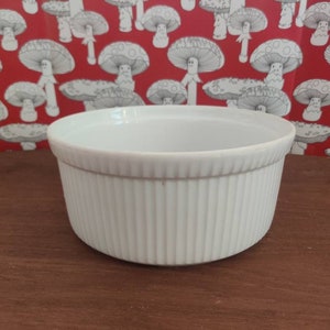 May include: A white ceramic ramekin with a circular shape and vertical fluted ridges along the exterior side. The bowl features a smooth, glossy finish and a wide, open top, providing a simple and functional design for various culinary uses.