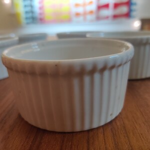 May include: A collection of white, ribbed ceramic ramekins. These small, round dishes have a classic design, perfect for baking individual portions of desserts or savory dishes. The ramekins are arranged on a wooden surface.