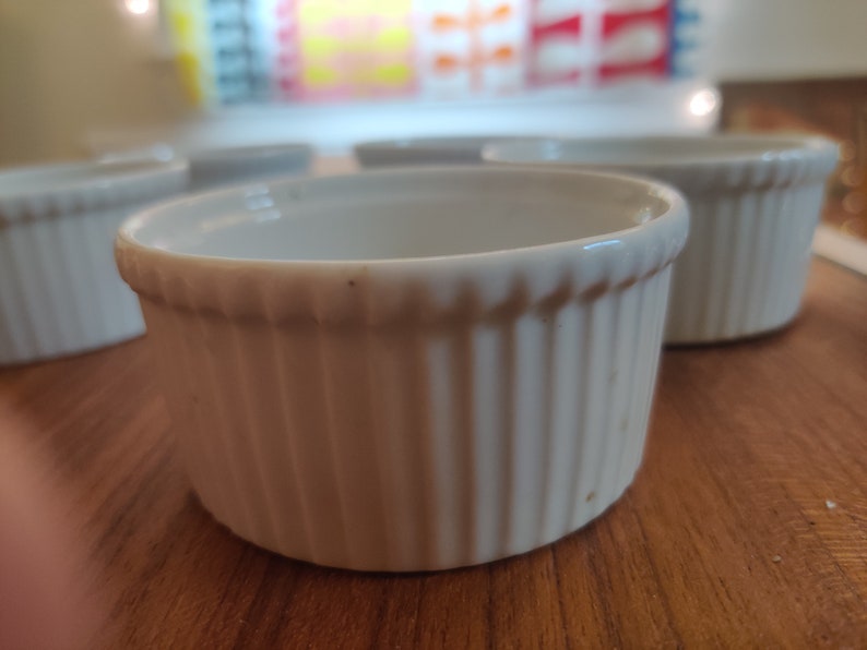May include: A collection of white, ribbed ceramic ramekins. These small, round dishes have a classic design, perfect for baking individual portions of desserts or savory dishes. The ramekins are arranged on a wooden surface.