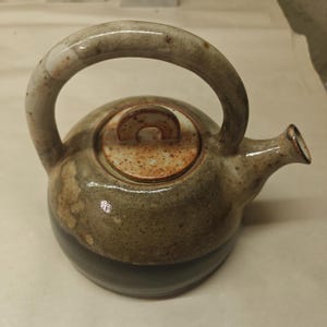 May include: A ceramic teapot with a rounded body, featuring a two-tone glaze with a dark charcoal base and a mottled, earthy green and tan top. It includes a curved handle, a short spout, and a matching circular lid with a small arched handle.
