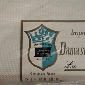 May include: A white fabric textile packaged in clear plastic with a label featuring a blue and white shield logo. The label text reads Warranted Damask, Cotton: 65%, Rayon: 35%, RN-27195, Cotton and Rayon, and lists dimensions as 58x88 and 8/15x15.