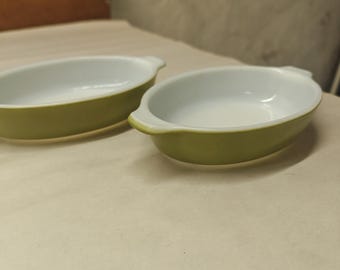 May include: Two oval-shaped baking dishes with a smooth, light green exterior and a glossy white interior. Each dish features small, integrated handles on opposite sides, providing a simple, functional design for oven-to-table serving.