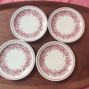 May include: A set of four round white ceramic plates featuring a decorative maroon floral pattern around the rim. Each plate has a smooth, circular center and a raised edge with a consistent maroon border, showcasing a detailed botanical design.
