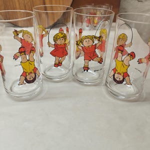 May include: A set of four clear glass tumblers featuring colorful Cabbage Patch Kids graphics. Each glass displays illustrations of children with yarn hair in red and yellow outfits, some jumping rope or on roller skates, with 1984 OAA text.