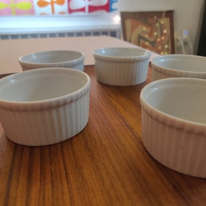 May include: Five white, ribbed ceramic ramekins are arranged on a wooden surface. The ramekins have a classic design with a slightly flared rim and are ideal for baking individual portions of desserts or savory dishes.