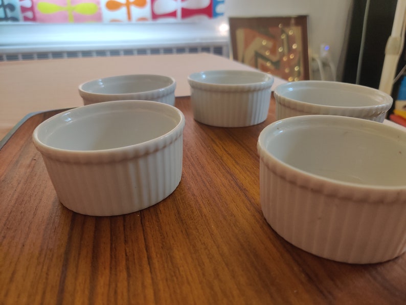 May include: Five white, ribbed ceramic ramekins are arranged on a wooden surface. The ramekins have a classic design with a slightly flared rim and are ideal for baking individual portions of desserts or savory dishes.
