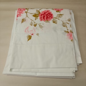 May include: A white pillowcase featuring a floral pattern of pink roses and green leaves across the top section. The fabric is smooth and opaque, with a clean hemmed edge visible at the bottom. The design is concentrated on the upper portion.