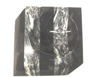 May include: A square-shaped ashtray crafted from polished black stone, featuring two prominent vertical bands of white and grey marble veining that run across the surface and through the central circular basin of the object.