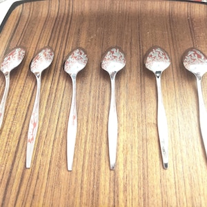 May include: Six vintage silver spoons with a decorative pattern on the spoon bowls are arranged on a wooden tray. The spoons have long, slender handles and are displayed against a warm, brown background.