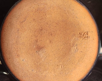 May include: A circular, unglazed terracotta-colored ceramic base with a speckled texture. The surface features a stamped maker's mark that reads ALFA OCHI in a rectangular border. The base is surrounded by the dark, glossy rims of other ceramic vessels.