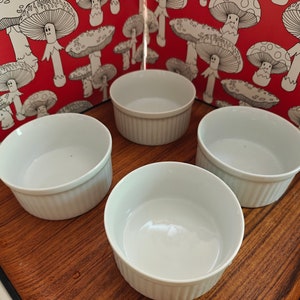 May include: Four white ceramic ramekins with vertical fluted sides arranged on a wooden tray. Each ramekin features a smooth interior and a consistent cylindrical shape, providing a clean and functional design for serving individual portions of food.