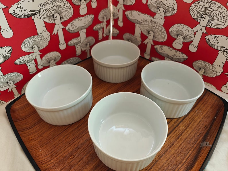May include: Four white ceramic ramekins with vertical fluted sides arranged on a wooden tray. Each ramekin features a smooth interior and a consistent cylindrical shape, providing a clean and functional design for serving individual portions of food.