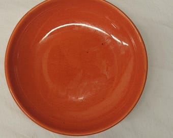 May include: A round, shallow ceramic bowl with a smooth, glossy finish in a vibrant, solid orange color. The bowl features a simple, circular design with a slightly raised rim and a uniform, consistent glaze across the entire interior surface.