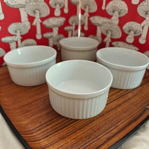 May include: Four white ceramic ramekins with vertical ribbed sides arranged in a group. Each small round dish features a smooth interior and a consistent cylindrical shape, designed for individual servings of food or baking applications.