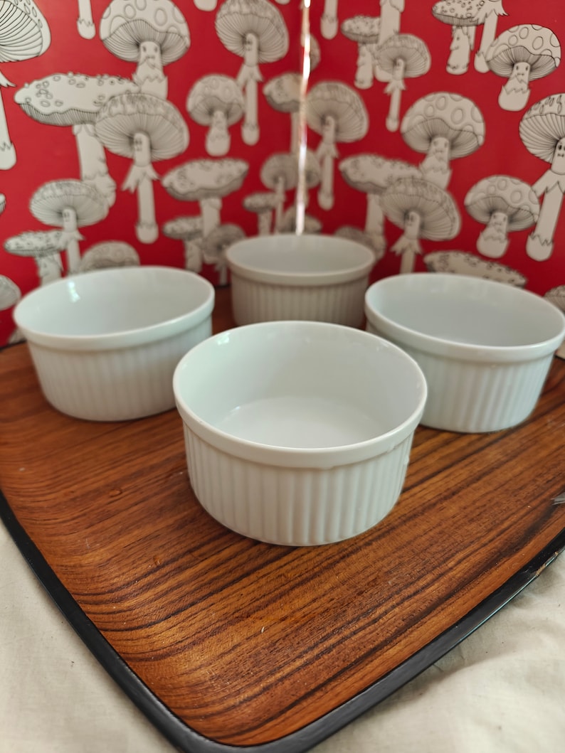 May include: Four white ceramic ramekins with vertical ribbed sides arranged in a group. Each small round dish features a smooth interior and a consistent cylindrical shape, designed for individual servings of food or baking applications.