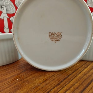 May include: A white ceramic ramekin with a fluted exterior and a red background featuring a white line-art mushroom pattern. The base is stamped with the text DANSK INTERNATIONAL DESIGNS LTD NR/JAPAN. The ramekin has a smooth, circular rim.