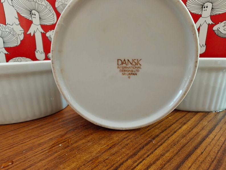 May include: A white ceramic ramekin with a fluted exterior and a red background featuring a white line-art mushroom pattern. The base is stamped with the text DANSK INTERNATIONAL DESIGNS LTD NR/JAPAN. The ramekin has a smooth, circular rim.
