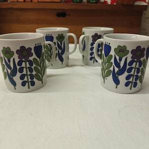 May include: A set of four white ceramic mugs, each featuring a repeating pattern of stylized flowers in shades of purple, blue, and green. The mugs have a cylindrical shape with a side handle and a smooth, glossy finish on the exterior surface.