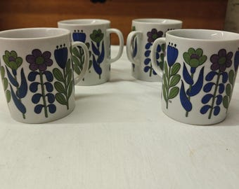 May include: A set of four white ceramic mugs featuring a repeating pattern of stylized flowers in shades of blue, purple, and green. Each mug has a handle on the side and a cylindrical shape with a smooth finish and a consistent floral design.