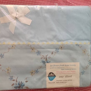 May include: A light blue bed sheet in clear plastic packaging with a white ribbon bow. The sheet features a floral bouquet pattern, a yellow scalloped trim, and a label reading "BOUQUET PATTERN" and "one sheet" with dimensions of 72 x 108 inches.