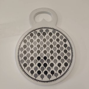 May include: A white, round trivet with a handle, featuring a pattern of gray and black stylized tree shapes. The edge is decorated with a black and white zigzag design. The trivet is designed to protect surfaces from hot items.
