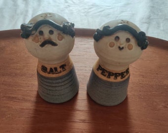May include: A pair of ceramic salt and pepper shakers featuring hand-painted faces with dark hair, rosy cheeks, and distinct expressions. The salt shaker has a mustache, while the pepper shaker has a small smile. Both have textured grey bases.