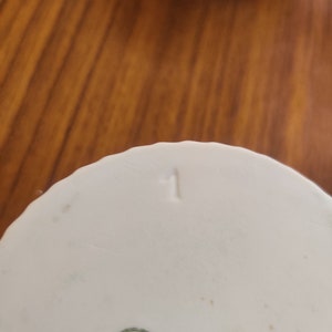 May include: Close-up of a white porcelain object with a ribbed design and the number "1" stamped on the bottom. The object has a small green and white label with text. The background is a wooden surface.