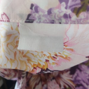 May include: Close-up of a floral print fabric featuring a collar. The fabric displays a pattern of pink, purple, and yellow flowers on a white background. A white label is attached to the collar.