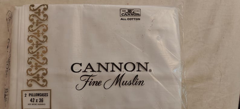 Może przedstawiać: Opakowanie białych poszewek na poduszki Cannon Fine Muslin. Opakowanie zawiera dwie poszewki na poduszki, każda o wymiarach 106,7 x 91,4 cm. Nazwa marki i szczeg&oacute;ły produktu są wydrukowane na czarno. W prawym g&oacute;rnym rogu znajduje się logo z napisem "Cannon All Cotton".