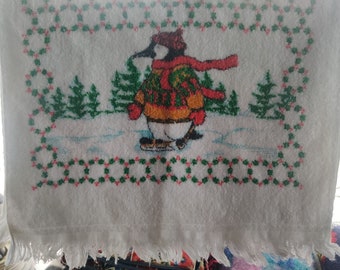 May include: A white rectangular hand towel featuring a fringed bottom edge and a central graphic of a penguin wearing a red hat, red mittens, and a colorful sweater while ice skating. The scene includes green pine trees and a red and green border.