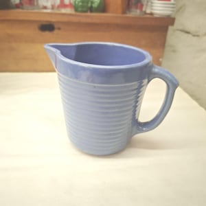May include: A periwinkle blue ceramic pitcher featuring a series of horizontal ribbed textures along the exterior body, a curved handle on the right side, and a small pouring spout at the top rim, finished with a smooth matte glaze throughout.