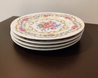 May include: A stack of four white ceramic plates featuring a central floral bouquet of pink, yellow, and blue flowers. The rim is decorated with a matching floral pattern and gold-colored filigree accents on a cream-colored background.