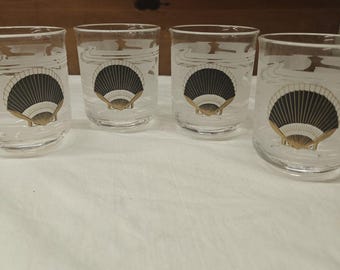 May include: A set of four clear glass tumblers, each featuring a central graphic of a black and gold fan-shaped seashell. The glasses have a smooth, cylindrical shape with a subtle frosted band design encircling the upper portion of the glass.