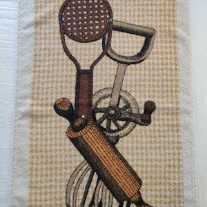 May include: Kitchen-themed towel featuring a brown rolling pin, whisk, and other cooking utensils. The background has a yellow and white checkered pattern. The towel has a white fringe along the top edge.