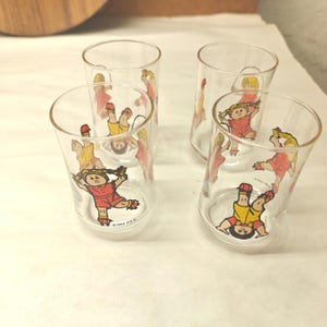 May include: A set of four clear glass tumblers, each decorated with colorful illustrations of Cabbage Patch Kids dolls in various active poses, including jumping rope and doing handstands, with the text 1984 O.A.A. printed on the glass.