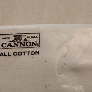 Może przedstawiać: Biały produkt tekstylny w przezroczystym plastikowym opakowaniu. Produkt posiada czarno-białą etykietę z napisem "MADE IN U.S.A. CANNON ALL COTTON". Opakowanie jest częściowo widoczne.