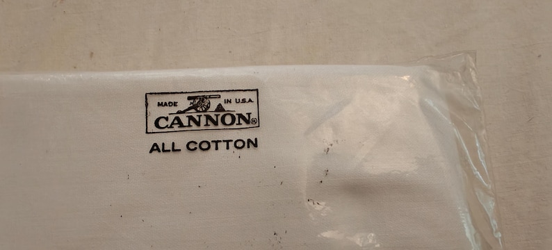 Może przedstawiać: Biały produkt tekstylny w przezroczystym plastikowym opakowaniu. Produkt posiada czarno-białą etykietę z napisem "MADE IN U.S.A. CANNON ALL COTTON". Opakowanie jest częściowo widoczne.