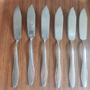 May include: Six stainless steel butter knives with a curved blade and ornate handle design, arranged on a wooden serving tray. The knives are reflective, catching the light, and are a classic addition to any dining set.