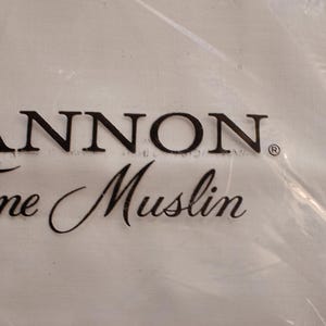 Może przedstawiać: Biała tkanina z czarnym napisem "CANNON. Fine Muslin". Tkanina znajduje się w przezroczystej plastikowej torbie, a tekst jest lekko zasłonięty przez plastik.