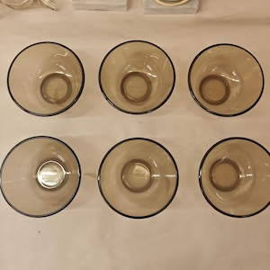 Peut inclure: Un ensemble de six bols ronds en verre assortis, teintés d'une couleur brun fumé translucide. Chaque bol présente un bord circulaire lisse et une base légèrement encastrée, disposés en deux rangées de trois. Le verre est uniforme.
