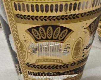May include: A clear glass tumbler featuring a decorative gold and black band around the upper portion. The design includes repeating geometric patterns, vertical lines, and stylized leaf motifs printed in metallic gold and solid black ink.