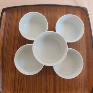 May include: Five small, white ceramic ramekins arranged on a rectangular wooden tray with a dark brown border. The ramekins are round with a simple, classic design, perfect for serving individual portions.