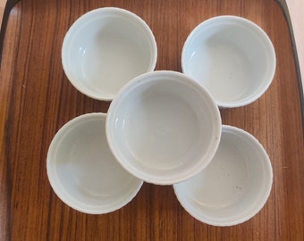 May include: Five small, white ceramic ramekins arranged on a rectangular wooden tray with a dark brown border. The ramekins are round with a simple, classic design, perfect for serving individual portions.