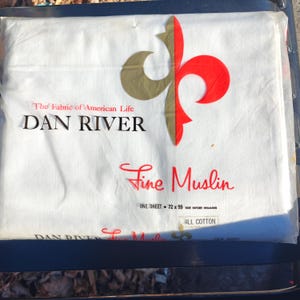 May include: A white Dan River fine muslin bed sheet in original packaging. The label features a gold and red fleur-de-lis logo, the text The Fabric of American Life, and states the size as 72 x 99 inches before hemming, made of all cotton.